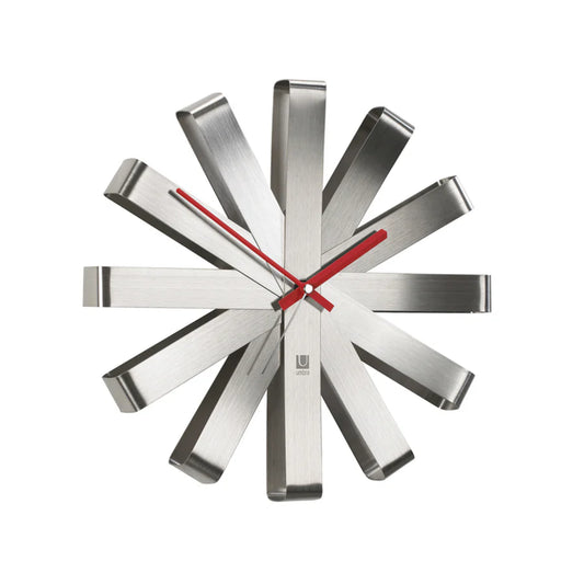 Relógio de parede UMBRA em metal prateado escovado, com design radial e ponteiros vermelhos, numa sala moderna sobre um aparador de madeira.